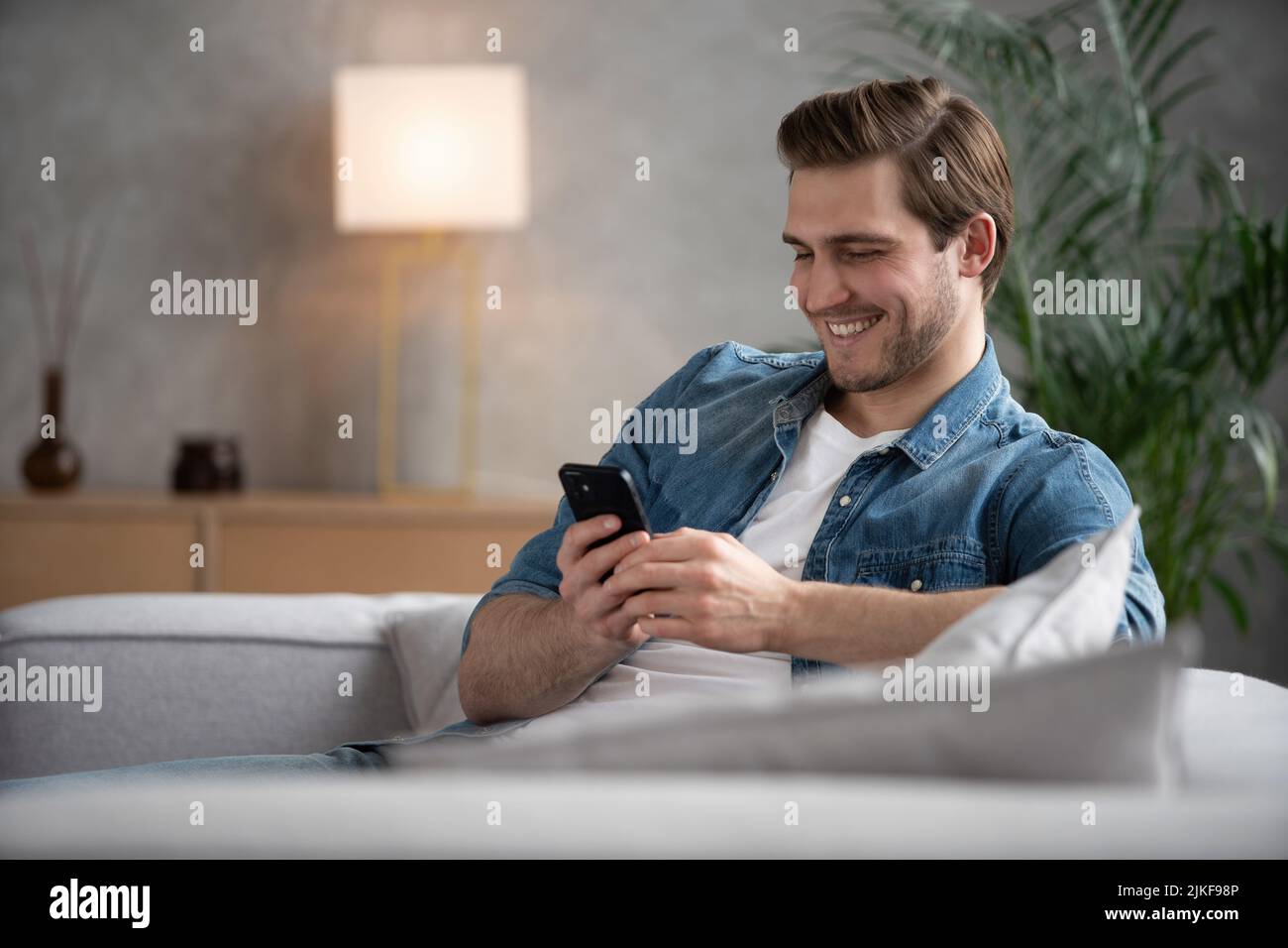 Attraktiver, lächelnder junger Mann in legerer Kleidung, der auf einer Couch im Wohnzimmer sitzt und ein Mobiltelefon benutzt Stockfoto