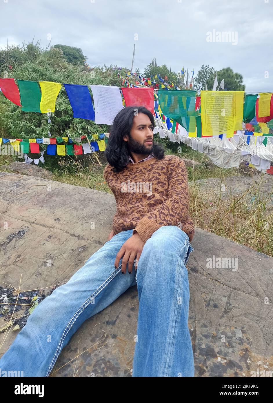 Ein attraktiver junger Mann aus Indien mit langen Haaren und Bart, der zur Seite schaut, während er auf Felsen sitzt und auf dem Hintergrund tibetischer Gebetsfahnen steht Stockfoto