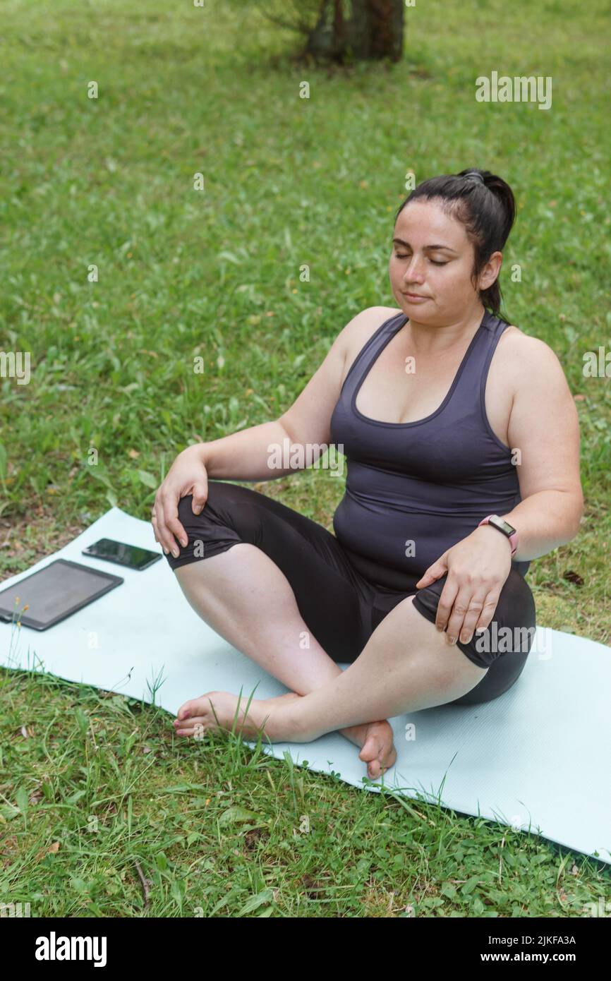 Eine charmante Brünette Frau plus-Größe Körper positiv praktiziert Sport in der Natur. Frau macht Yoga und Meditation im Park auf einer Sportmatte Stockfoto