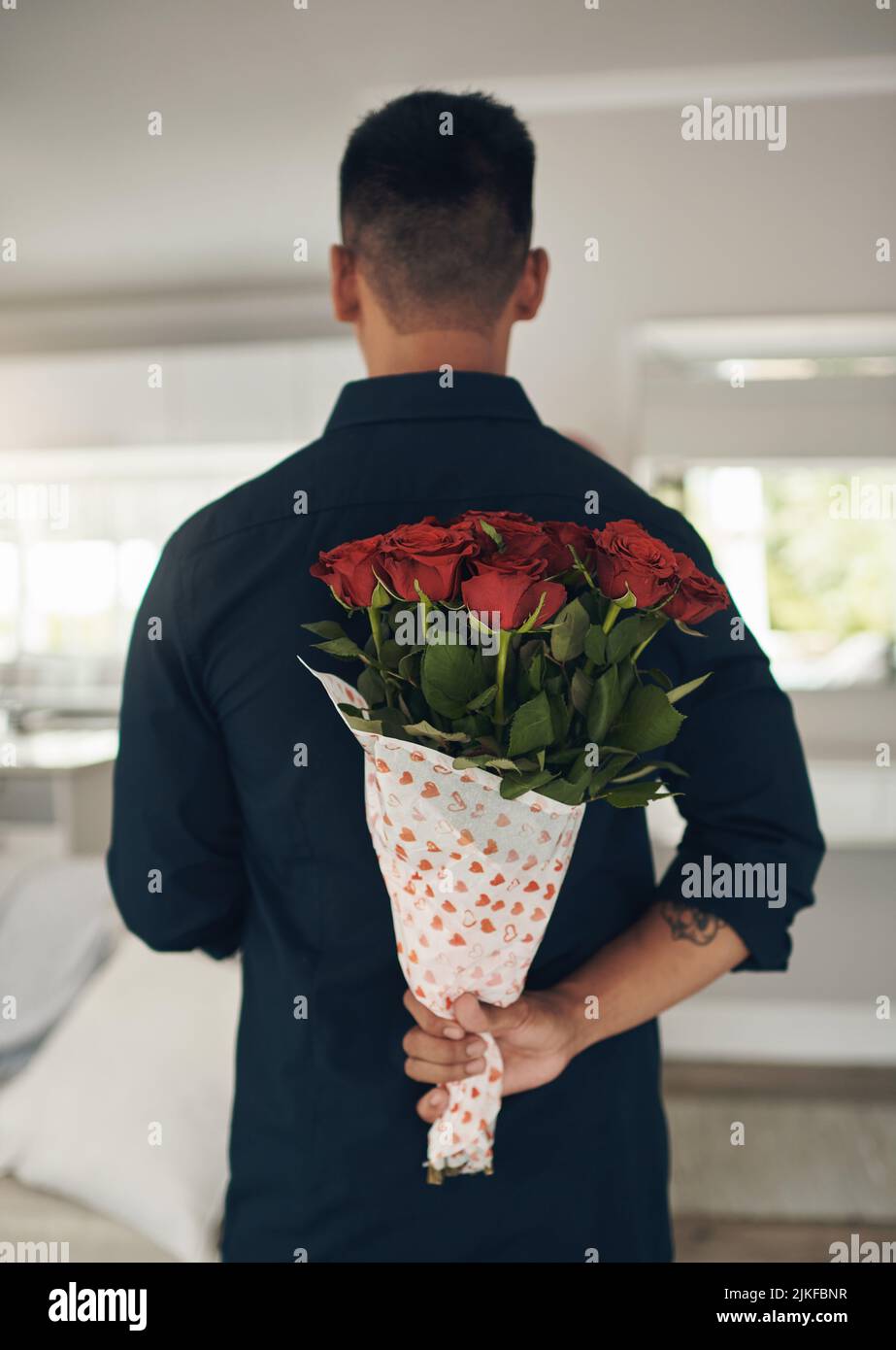 Kannst du erraten, was hinter meinem Rücken ist? Ein junger Mann, der zu Hause Blumen hinter seinem Rücken hält. Stockfoto