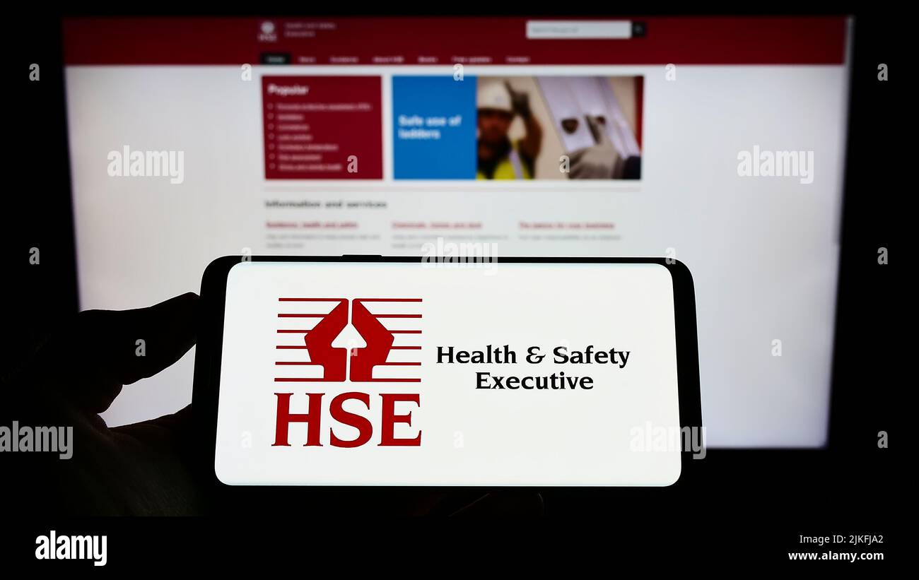 Person mit Mobiltelefon und Logo der britischen Agentur Health and Safety Executive (HSE) auf dem Bildschirm vor der Webseite. Konzentrieren Sie sich auf die Telefonanzeige. Stockfoto