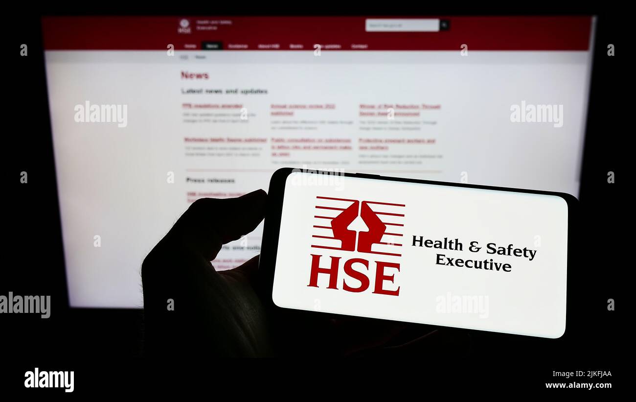 Person mit Mobiltelefon und Logo der britischen Agentur Health and Safety Executive (HSE) auf dem Bildschirm vor der Webseite. Konzentrieren Sie sich auf die Telefonanzeige. Stockfoto