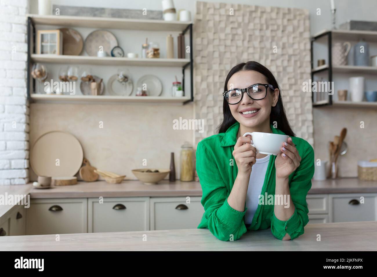 Junge schöne Frau in Brille und einem grünen Casual Shirt, zu Hause in der Küche, schaut auf die Webcam, Videoanruf hört auf den Gesprächspartner, Blick auf die Webcam Stockfoto