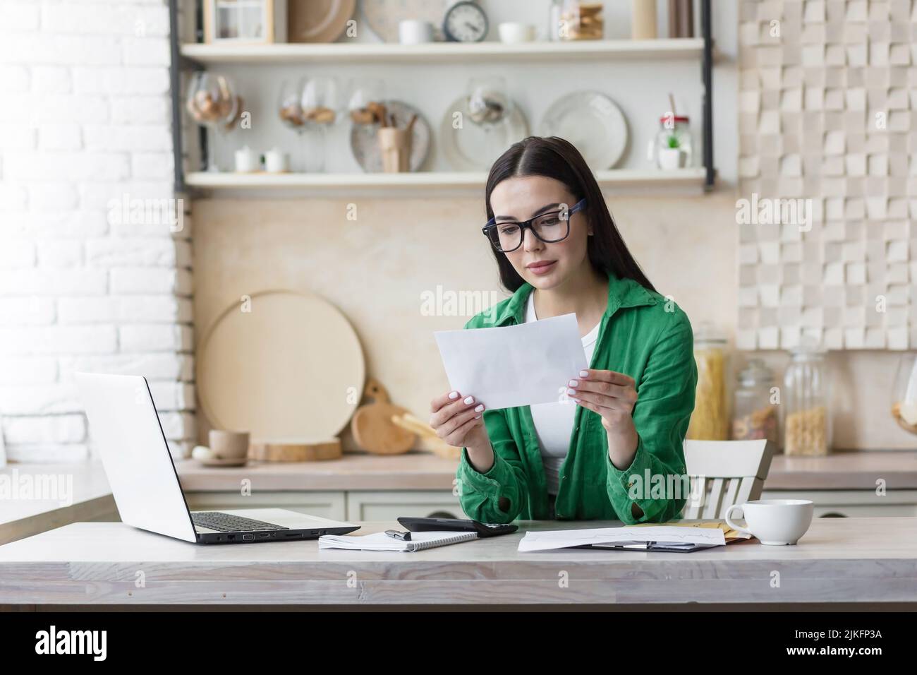 Seriöse und fokussierte Geschäftsfrau freiberuflich auf Papier arbeiten zu Hause in der Küche, brünett in der Brille und grün in der Küche, zahlen und Rechnungen zu berechnen Stockfoto