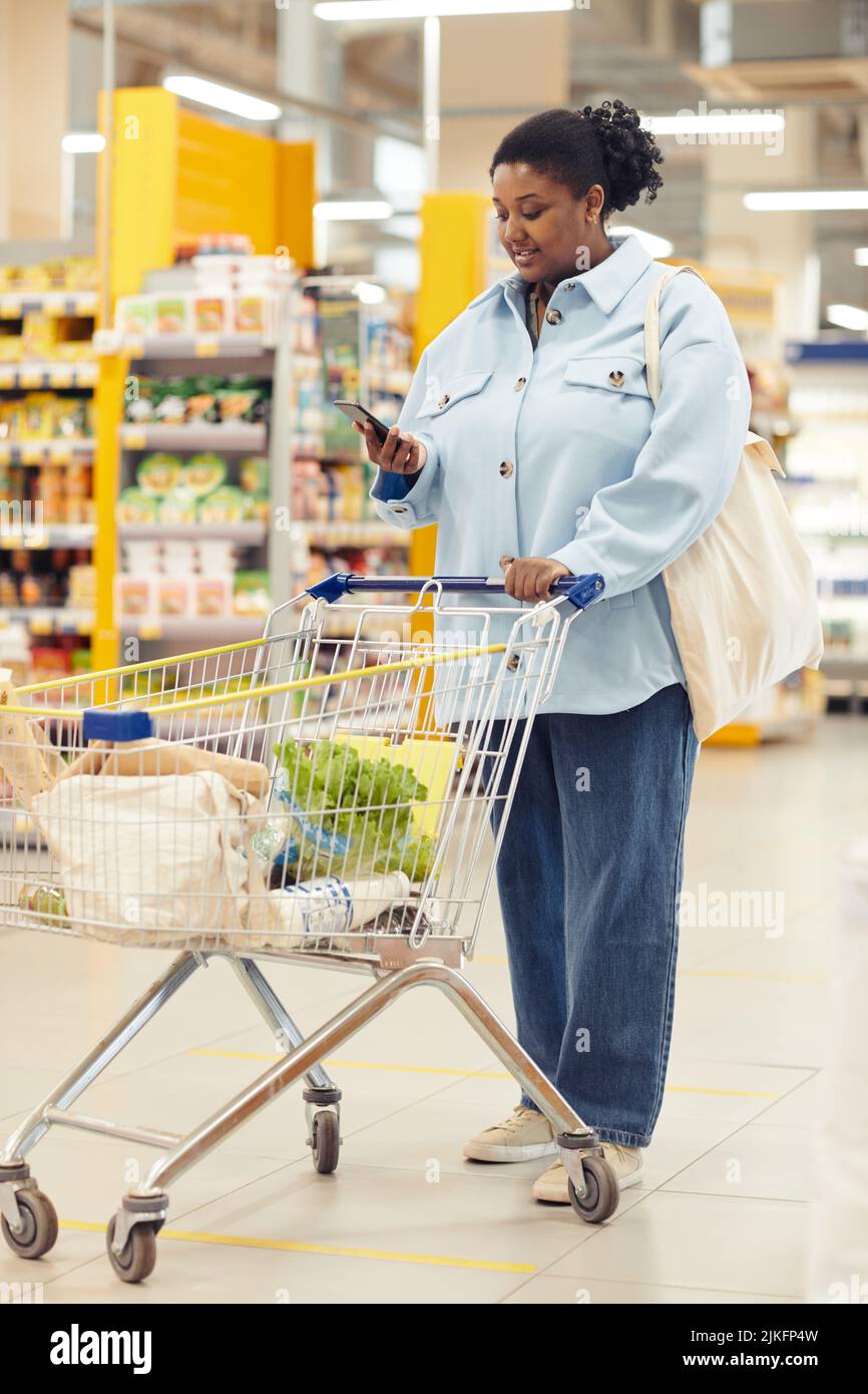 Vertikales Porträt einer schwarzen Frau, die Lebensmittel im Supermarkt kauft und das Smartphone benutzt Stockfoto