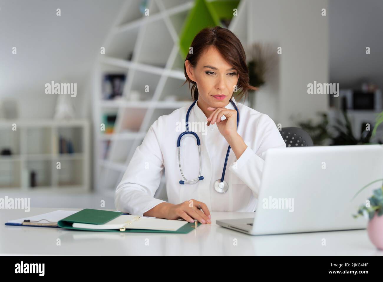 Aufnahme eines denkenden weiblichen Arztes, der hinter einem Laptop sitzt und in der Arztpraxis arbeitet. Stockfoto