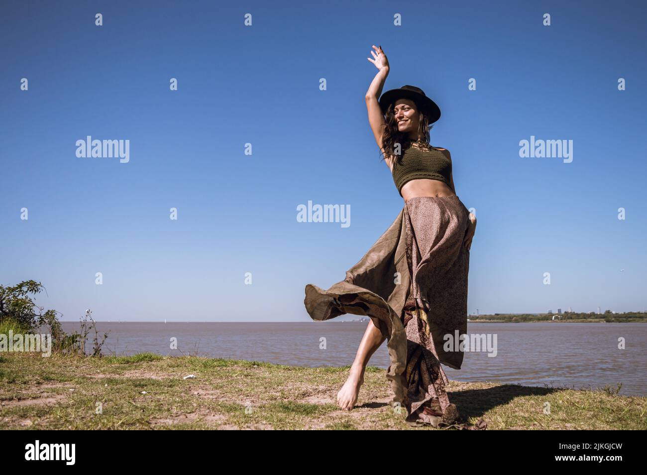 Die junge lateinische kaukasische Touristin, die ein langes Kleid und einen schwarzen Hut trägt, tanzt am Rand eines Gesimtes am Ufer des rio de la plata in ar Stockfoto