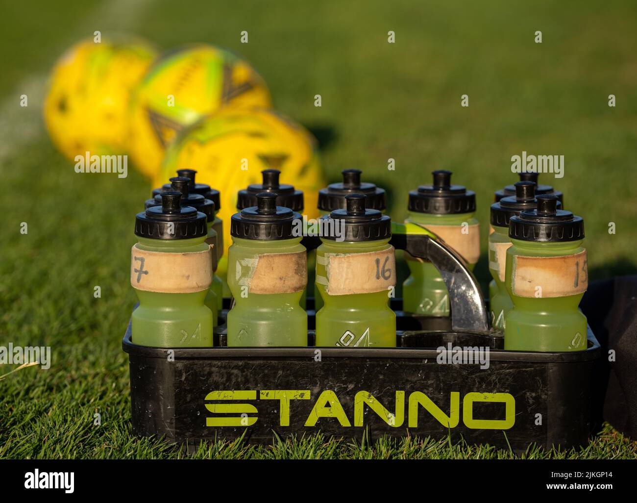 Gelbe, nachfüllbare Plastikflaschen mit schwarzen Tops sitzen in einem STANNO-Träger auf einem Grasfußballplatz (Fußball). Drei gelbe Kugeln aus dem Fokus Stockfoto