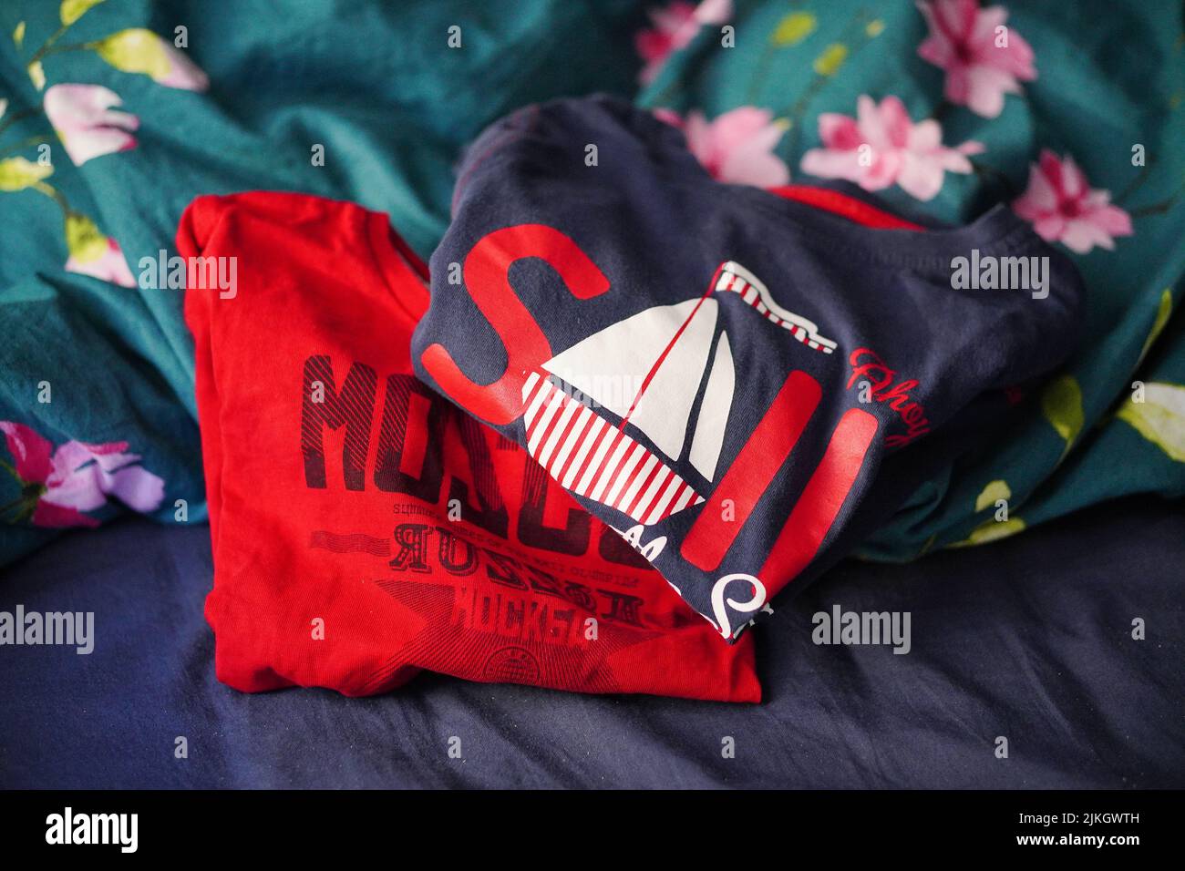 Ein rot-blaues Kinderhemd mit dem Text Sail and Moscow, das auf einem Bettlaken mit Blumen liegt Stockfoto