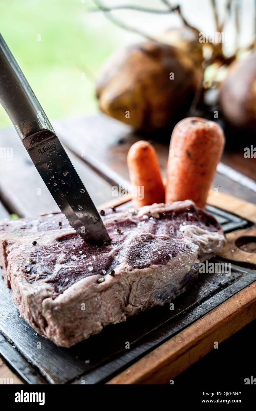 Eine vertikale Nahaufnahme eines Messers in einem rohen Steak mit Karotten auf einem Schneidebrett auf einem Holztisch Stockfoto