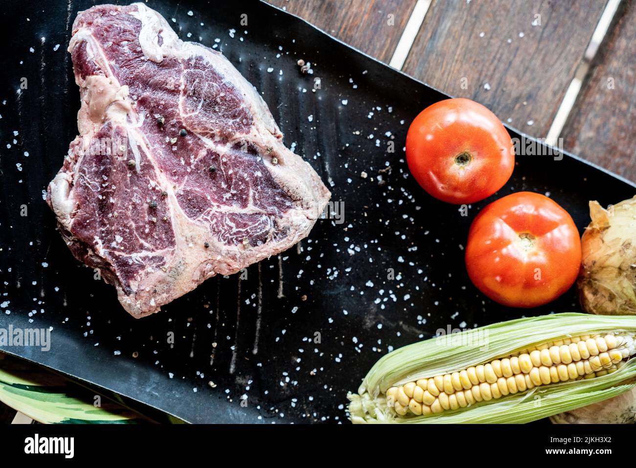 Ein Blick von oben auf ein rohes Steak mit Tomaten und Mais auf einem Brett auf einem Holztisch Stockfoto