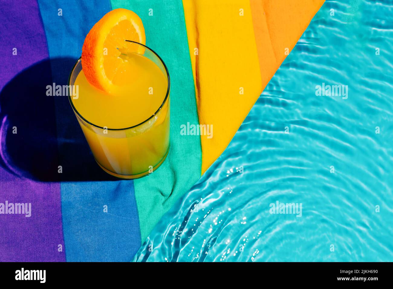Eine Tasse Orangensaft aus einem hohen Winkel mit orangefarbener Scheibe neben einem farbenfrohen Tuch am Pool Stockfoto