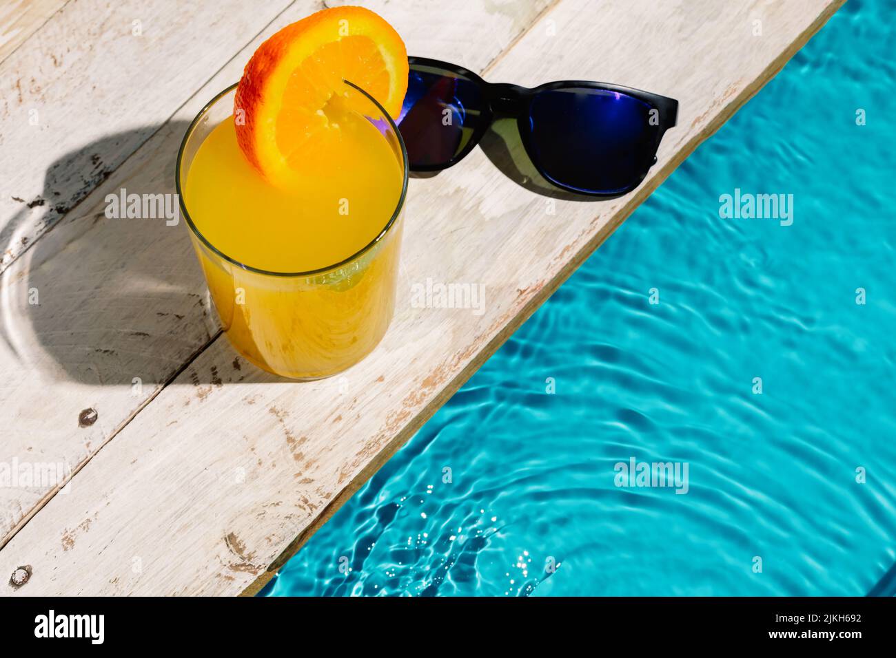 Eine Aufnahme einer Tasse Orangensaft mit Orangenscheibe und Sonnenbrille in einem hohen Winkel auf dem Pool Stockfoto