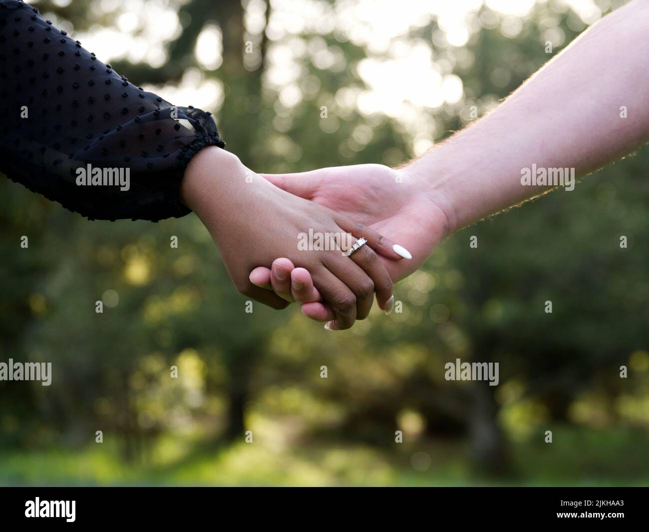 Ein schönes Paar, das sich vor einem verschwommenen Hintergrund die Hände im Freien an ihrer Verlobung hält Stockfoto