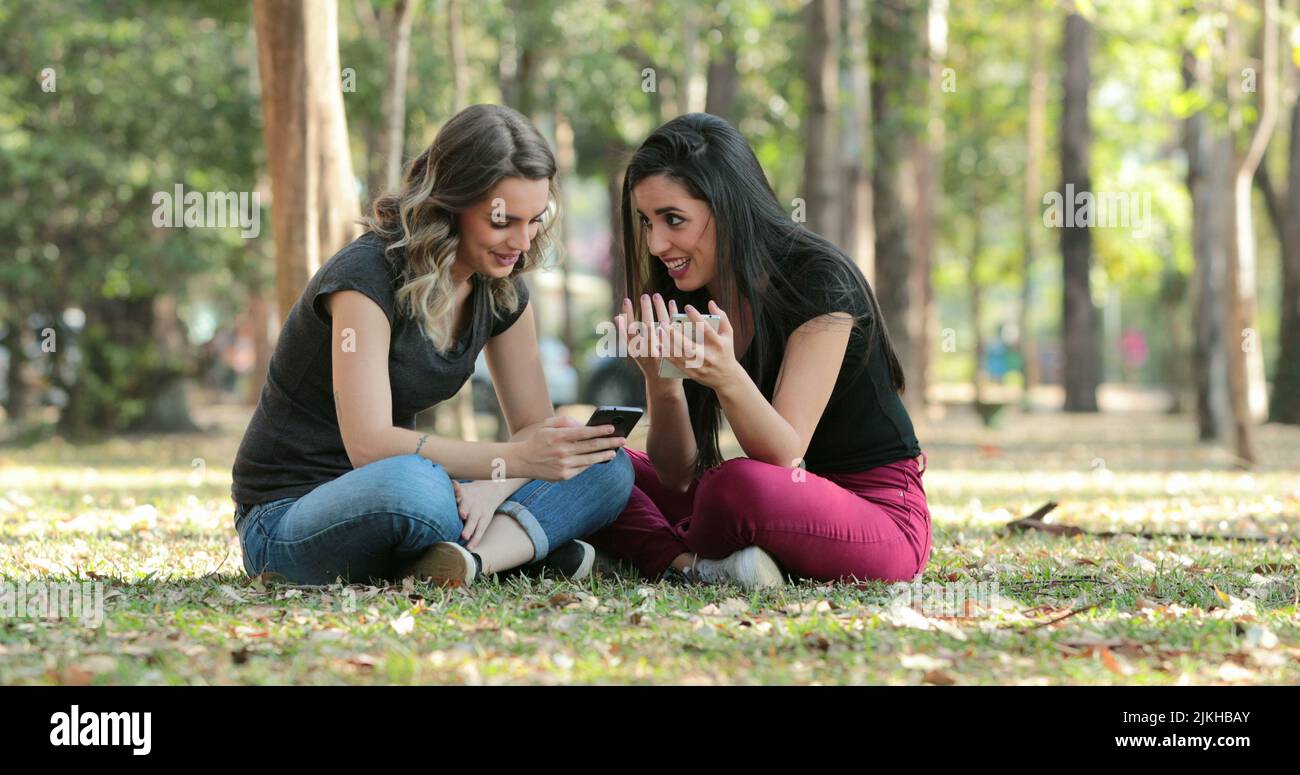 Ehrliche Freunde draußen im Park, die ihre Handys checken Mädchen sitzen und ihre Smartphones im Chat betrachten Stockfoto