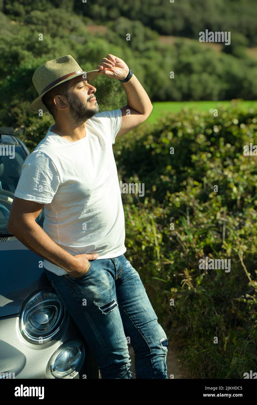 Ein glücklicher Mann, der in Jeans, weißem T-Shirt und mit der Hand einen Hut hält, genießt einen Frühlingstag in der Natur mit der Wärme der Sonne auf seiner Haut. Straße t Stockfoto