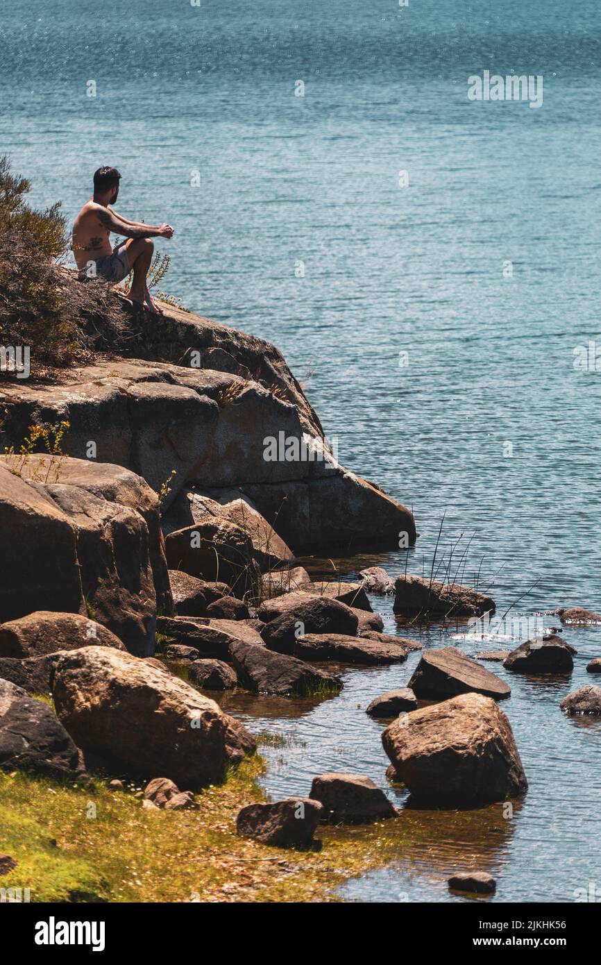 Ein junger Kaukasusmann, der auf dem Felsen sitzt und auf das Meer blickt Stockfoto