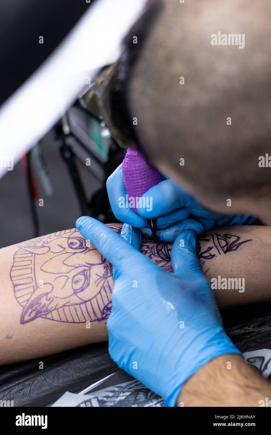 Vertikale Nahaufnahme eines alternativen Tätowierers, der in seinem kleinen Tattoo-Studio die Haut eines Kunden mit blauen Schutzhandschuhen tätowiert Stockfoto
