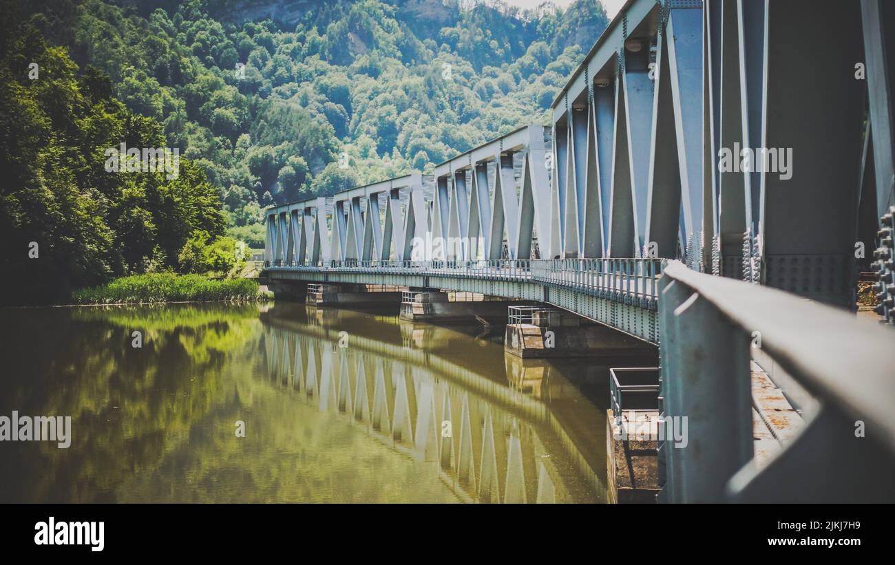 Eine metallische Fußbrücke in der Nähe des Flusses mit der Reflexion im Wasser in einem Dorf an einem sonnigen Tag Stockfoto