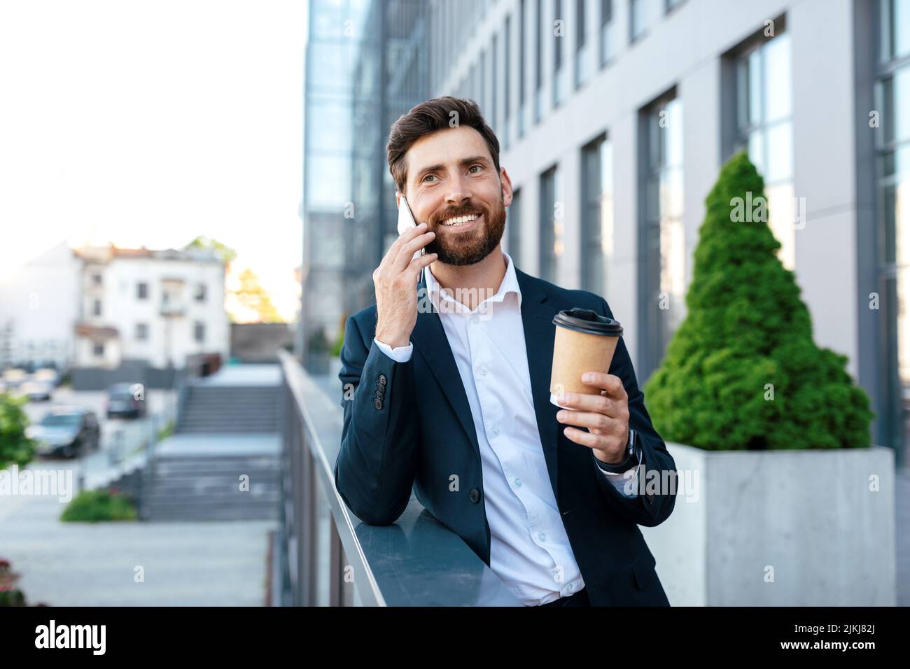 Glücklicher Tausendjähriger europäischer Rüde mit Bart im Anzug per Smartphone, Getränke vom Tassenbecher auf dem Balkon Stockfoto