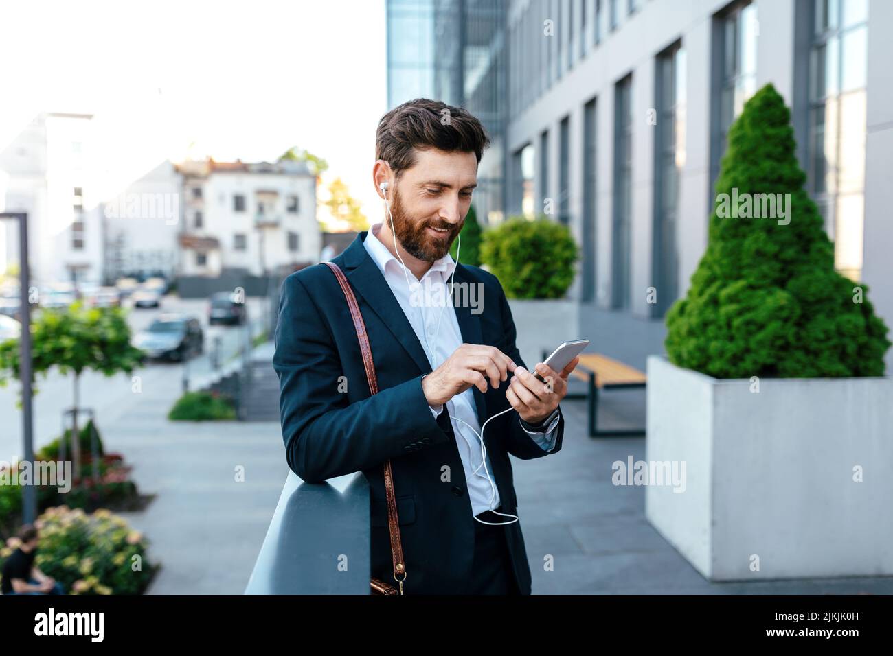 Lächelnder, tausendjähriger kaukasischer Geschäftsmann mit Bart im Anzug und Kopfhörern, die am Telefon tippen und Musik hören Stockfoto