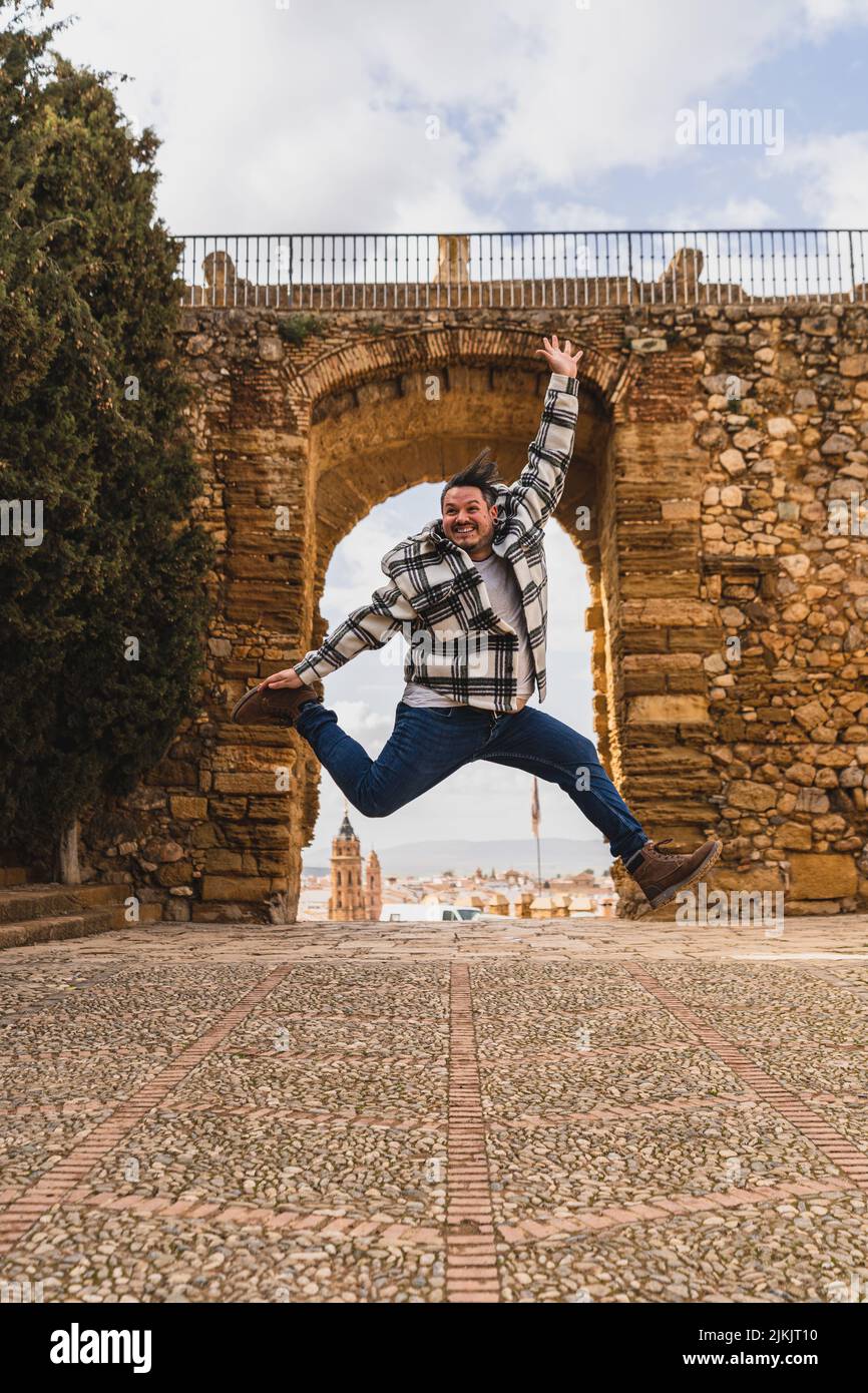 Eine vertikale Aufnahme eines jungen Mannes in einem karierten Hemd, der vor Freude in der Nähe einer historischen Brücke springt Stockfoto