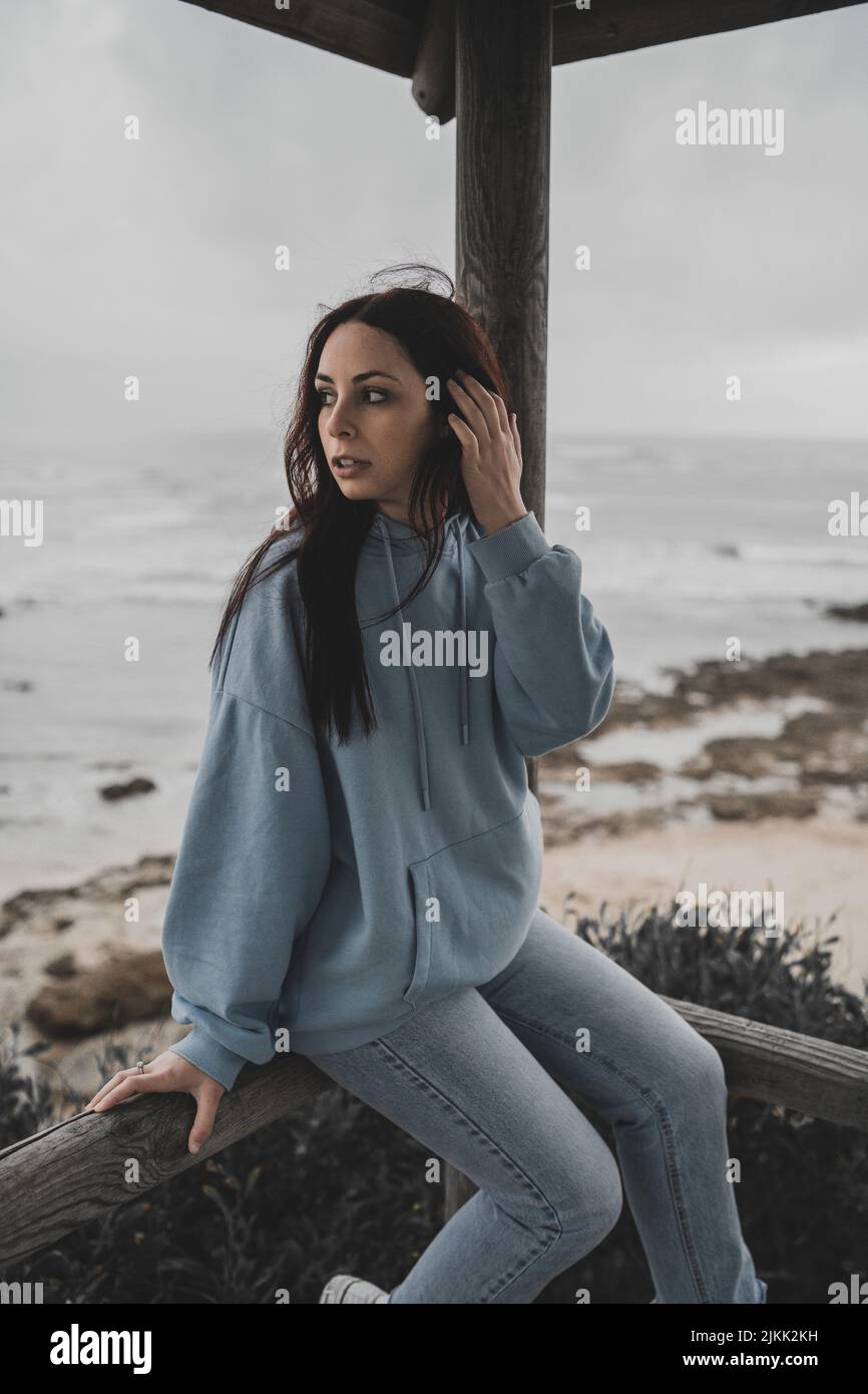 Eine niedliche junge kaukasische Dame mit Kapuzenpullover und Jeans sitzt in einem Pavillon am Meer Stockfoto