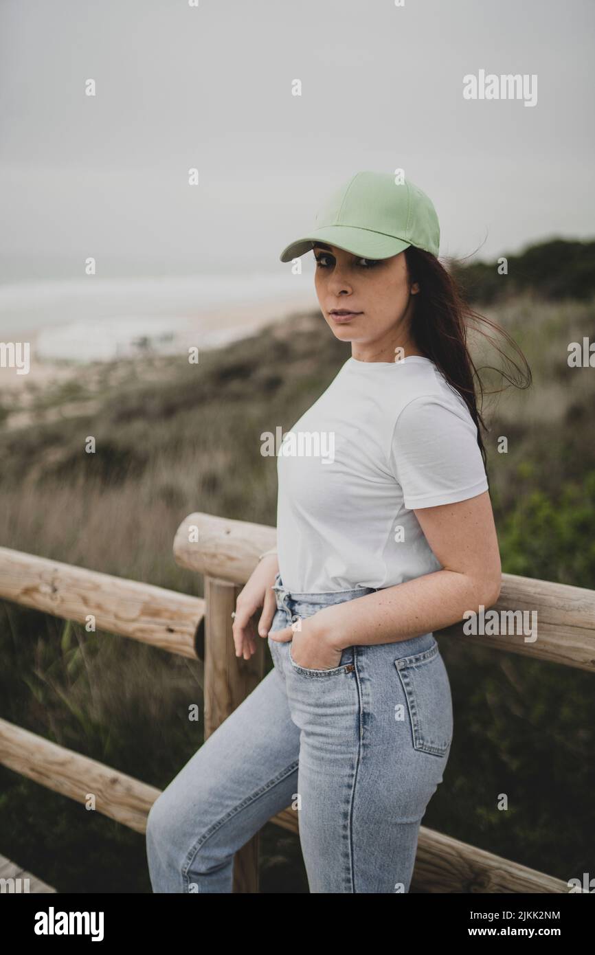 Eine niedliche junge kaukasische Dame, die eine Mütze, ein T-Shirt und eine Jeans trägt und in einem ländlichen Gebiet posiert Stockfoto