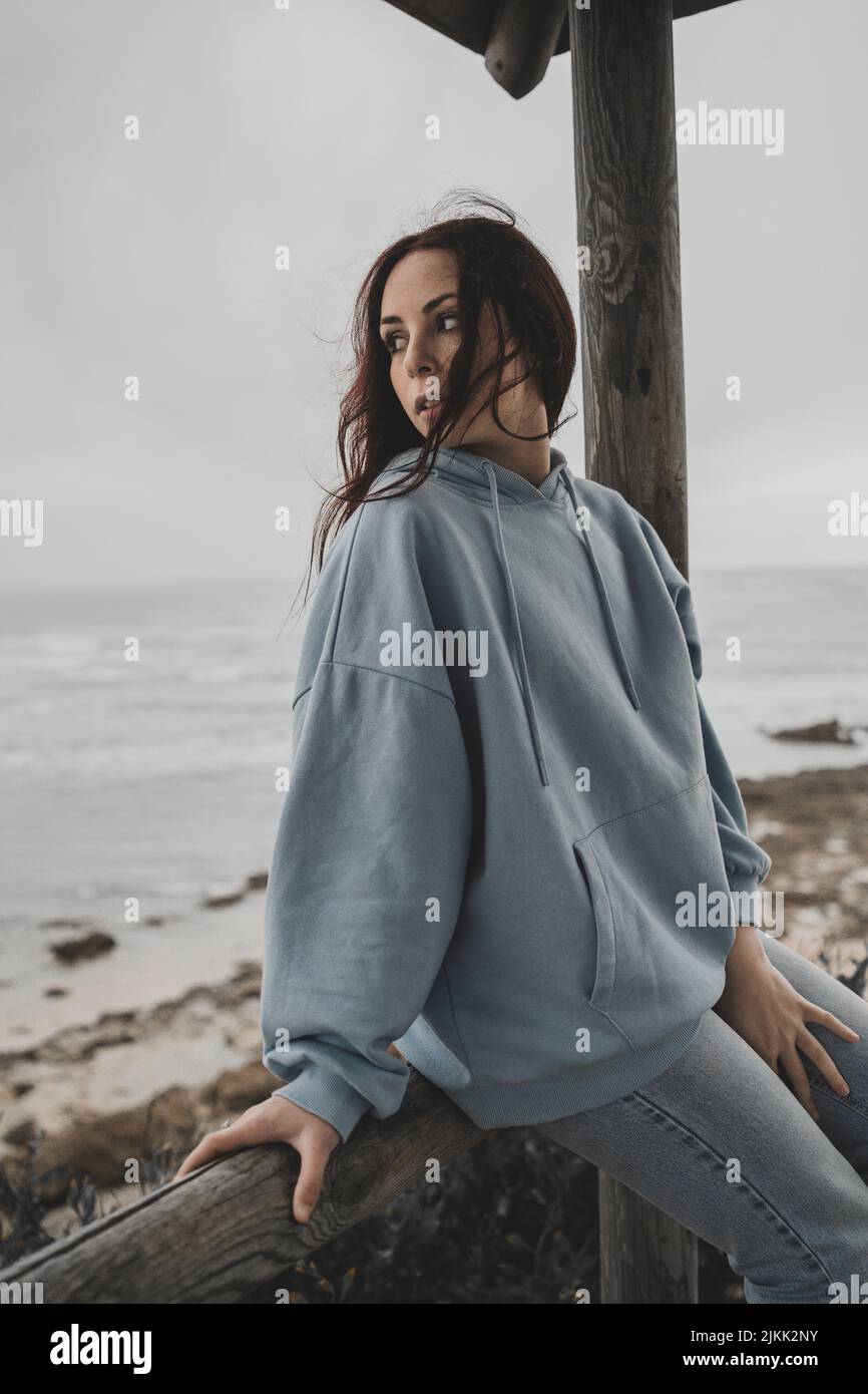 Eine niedliche junge kaukasische Dame mit Kapuzenpullover und Jeans sitzt in einem Pavillon am Meer Stockfoto
