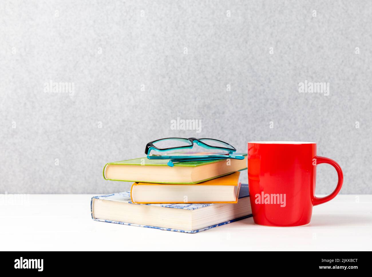 Eine Nahaufnahme einer Brille auf Büchern daneben und einer roten Tasse auf einem grauen Hintergrund und einem Kopierraum Stockfoto