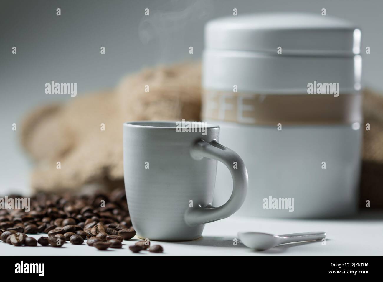 Ein flacher Fokus einer Tasse heißen Kaffees mit Kaffeebohnen und einem weißen Behälter im Hintergrund Stockfoto