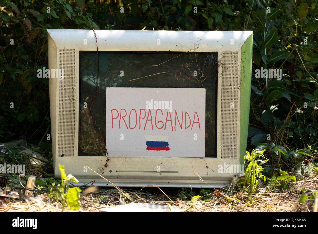 Ein alter zerbrochener Fernseher und Papppapier mit Textpropaganda und einer bemalten russischen Flagge und der Fernseher selbst steht im Wald auf dem Gras in der Ukraine. Stockfoto
