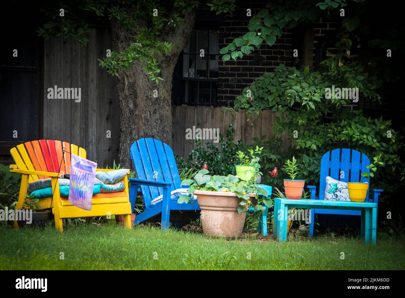 Farbenfroher Sitzbereich im Freien - bemalte Adirondack-Stühle mit Blumentöpfen unter Bäumen im schattigen Bereich und der Flagge von Home Sweet Home Stockfoto