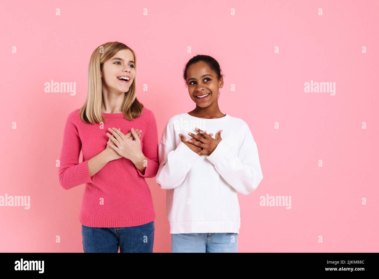 Multirassische zwei Mädchen lächeln, während sie die Hände auf ihre Brust halten, isoliert über die rosa Wand Stockfoto