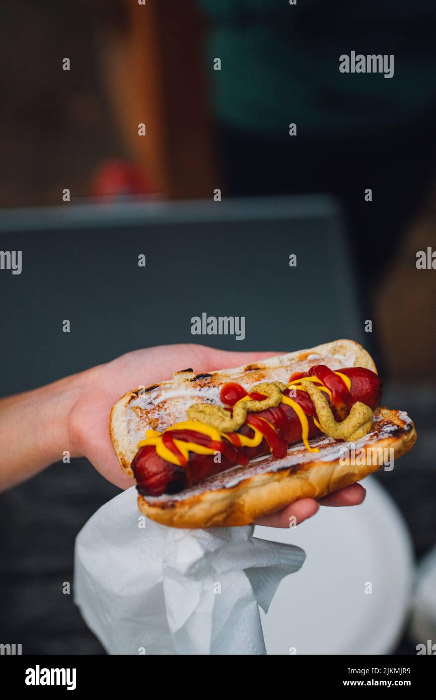 Eine vertikale Aufnahme einer Hand, die einen Hot Dog mit Ketchup, Senf und einer Papierserviette im Freien hält Stockfoto