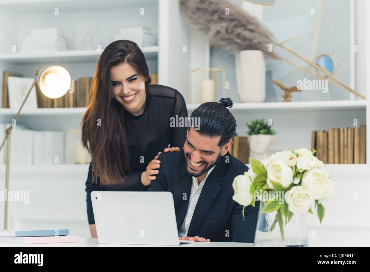 Die Freundin spielt sich spielerisch mit dem von zu Hause arbeitenden Geschäftsmann, der vor dem Laptop am Schreibtisch sitzt. Lachendes Paar. Innenaufnahme. Hochwertige Fotos Stockfoto