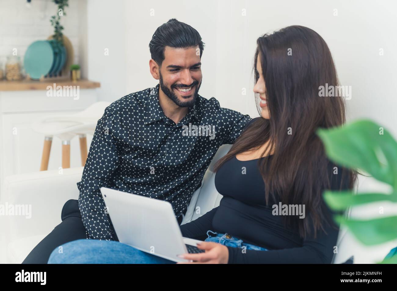 Ein Mann mit dunklem Haar und lächelndem Bart, der seiner Frau beim Arbeiten auf dem Laptop auf der Couch zuschaute. Junges Paar. Innenaufnahme. Hochwertige Fotos Stockfoto