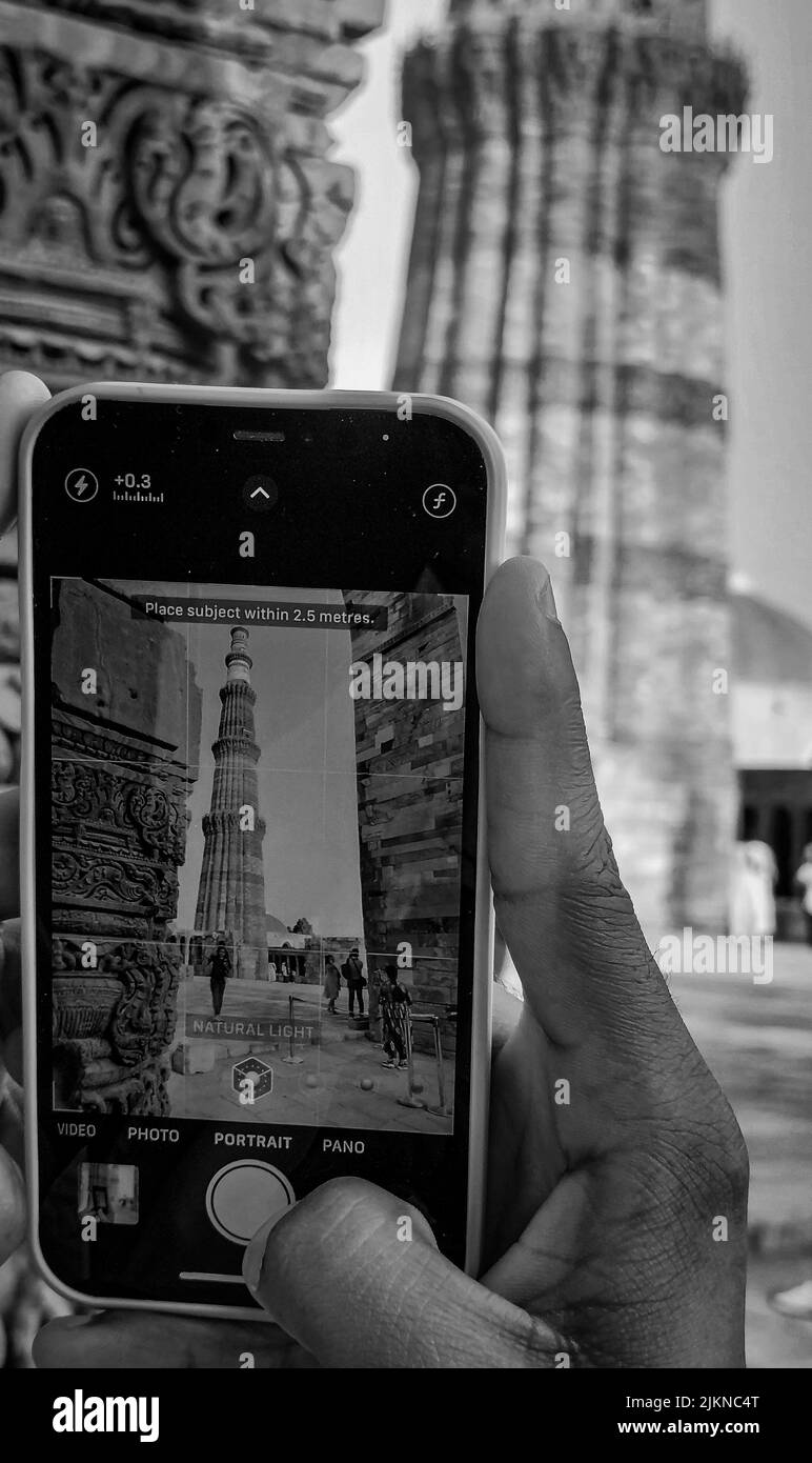 Eine Graustufe der Hand einer Person mit einem Telefon, das ein Foto eines alten Qutab Minar Turms macht Stockfoto