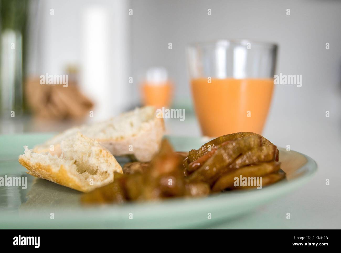 Nahaufnahme einer grünen Platte mit einem Stück Brot und Scheiben natürlicher Tomaten neben einem Glas cremigem Salmorejo auf einem Küchentisch. Gastronomischer Genuss Stockfoto