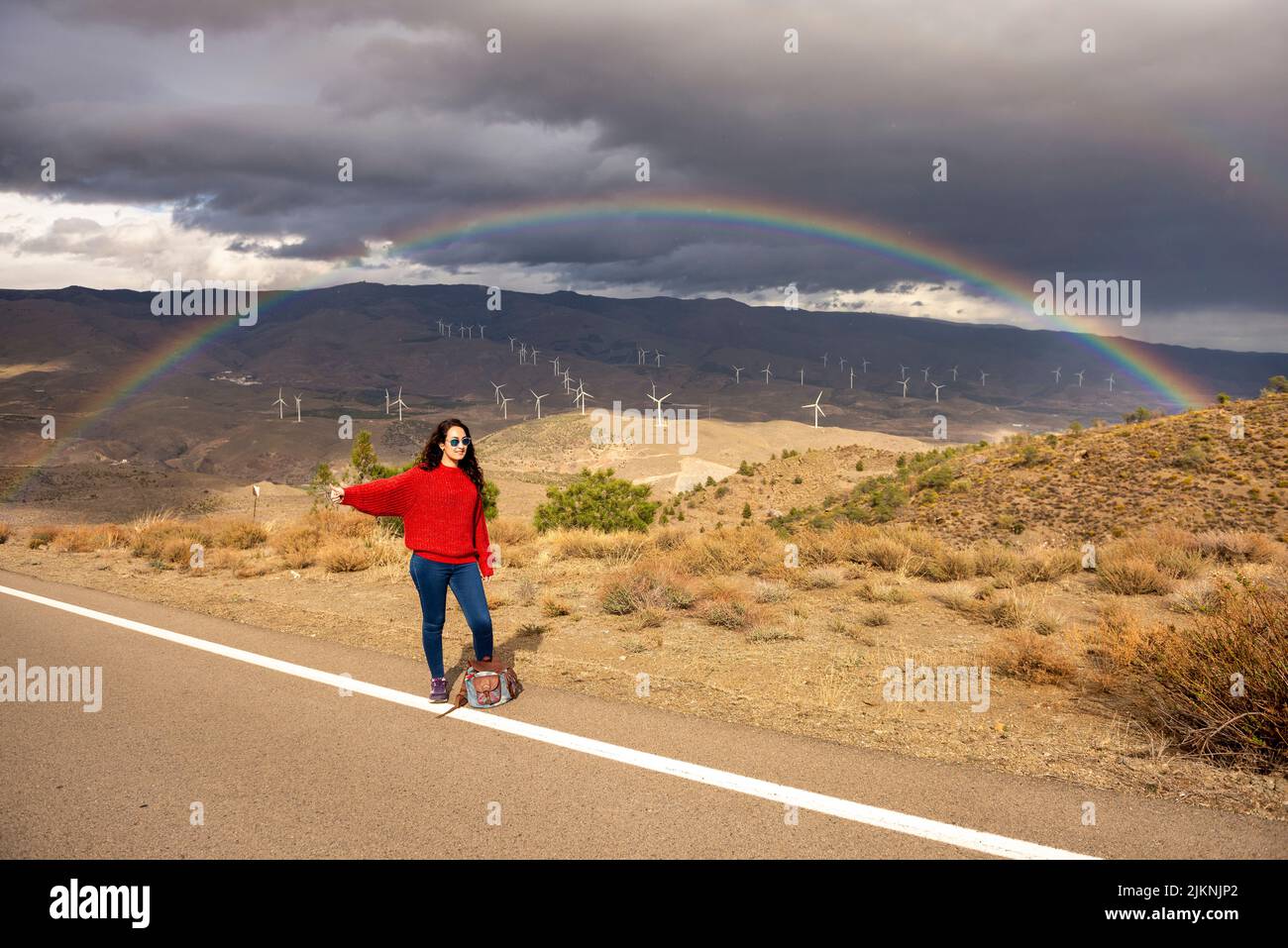 Frau in ihrem 25s Anhalter an einem Tag mit Sonne, Wolken und Regenbögen im Hintergrund. Stockfoto