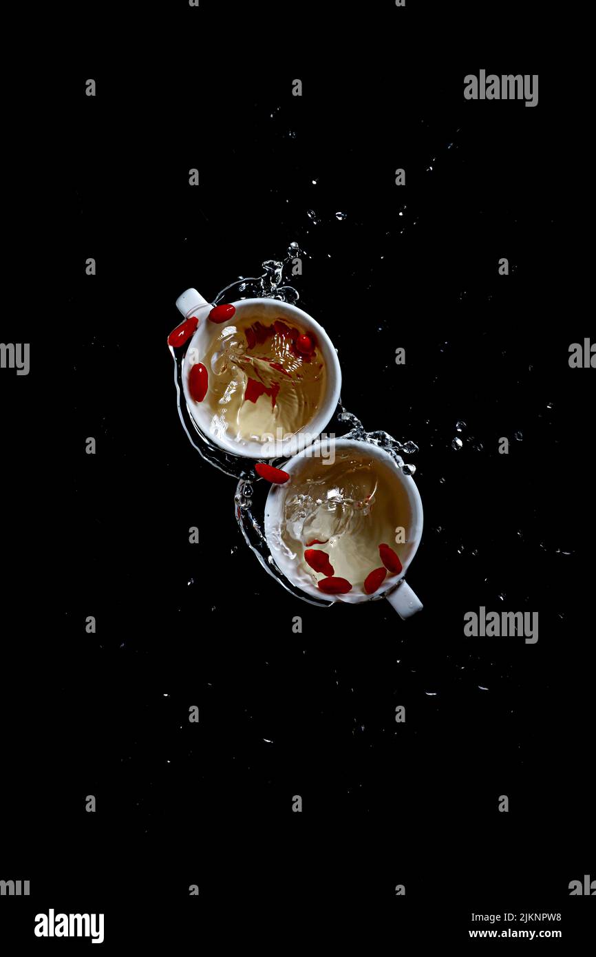 Eine Draufsicht auf zwei weiße Tassen mit spritzendem Wasser und roten Eckkörnern auf schwarzem Hintergrund Stockfoto