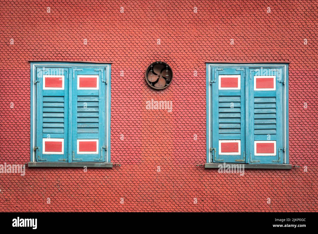 Zwei bemalte Fensterläden und ein Ventilator an einer orangefarbenen Wand eines Gebäudes Stockfoto