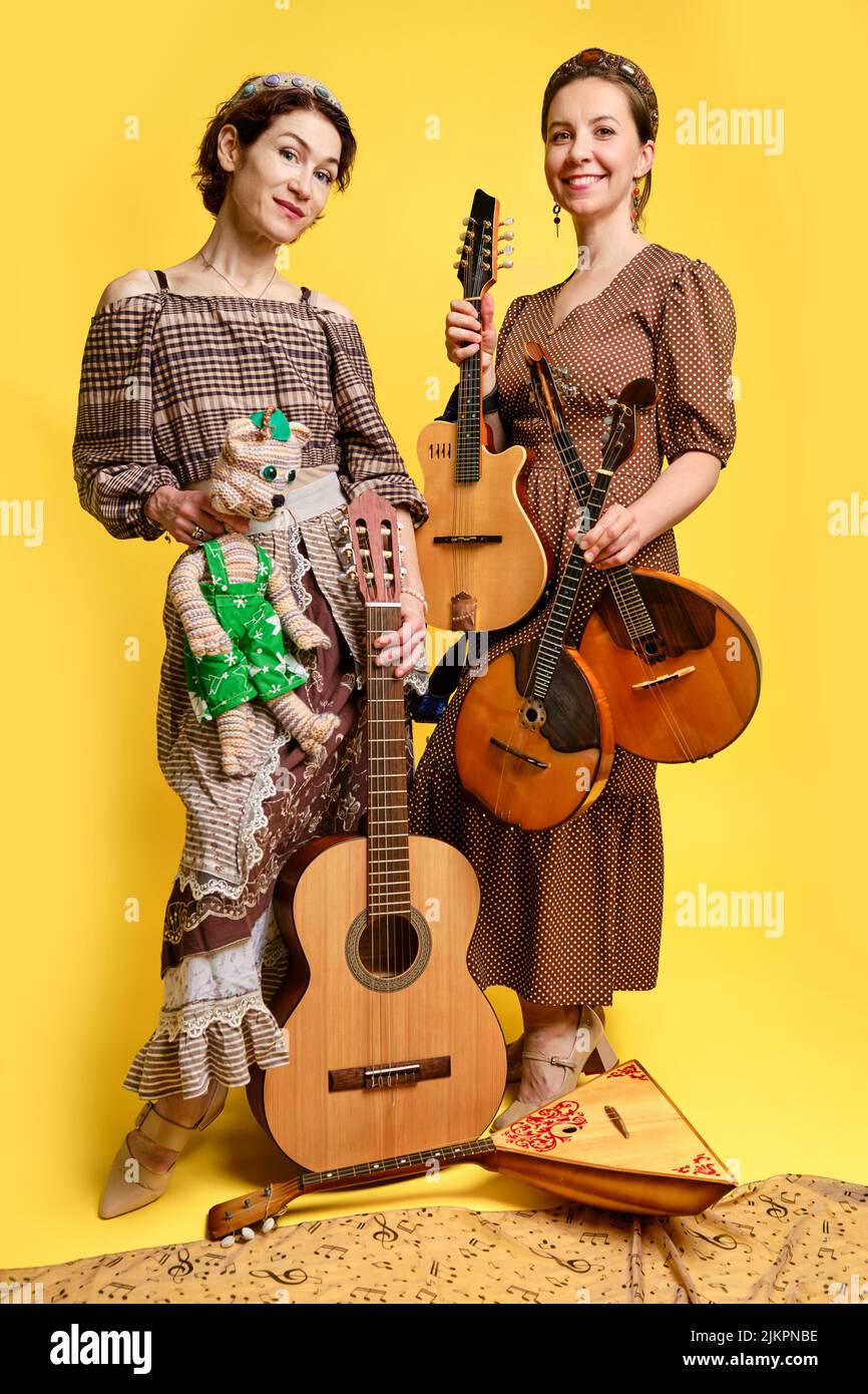 Musikerinnen in Kleidern mit Musikinstrumenten auf gelbem Studiohintergrund. Fröhliche Künstler mit Streichinstrumenten und einem Lächeln im Gesicht Stockfoto