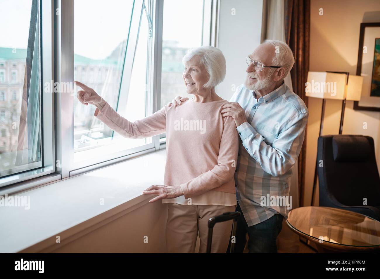 Das ältere Paar steht neben dem Fenster und sieht glücklich aus Stockfoto