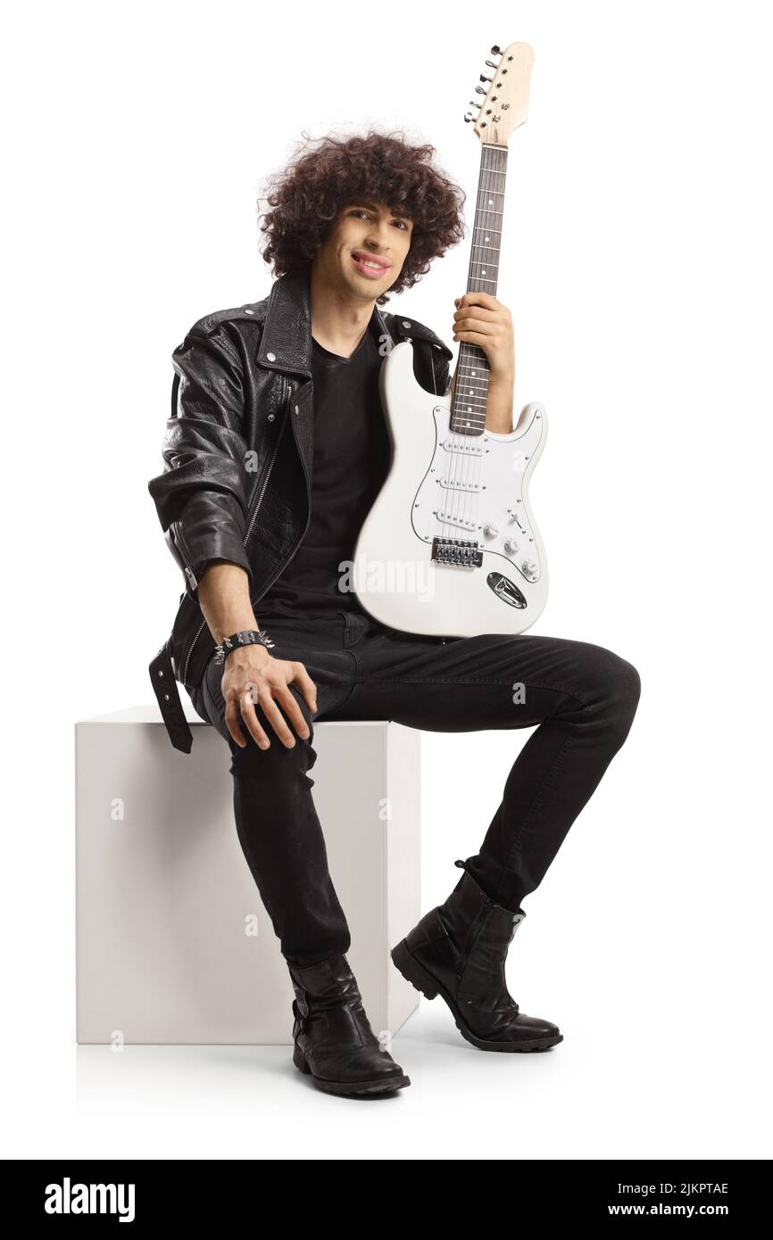 Lächelnder junger Rockmusiker, der eine Gitarre hält und auf einem weißen Würfel sitzt, der auf weißem Hintergrund isoliert ist Stockfoto