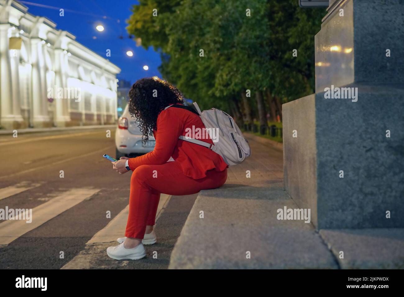 Eine dicke Frau mit langen, lockigen dunklen Haaren in einem leuchtend roten Anzug mit einem Telefon in den Händen sitzt im Zentrum einer europäischen Stadt bei einem cro für Fußgänger Stockfoto
