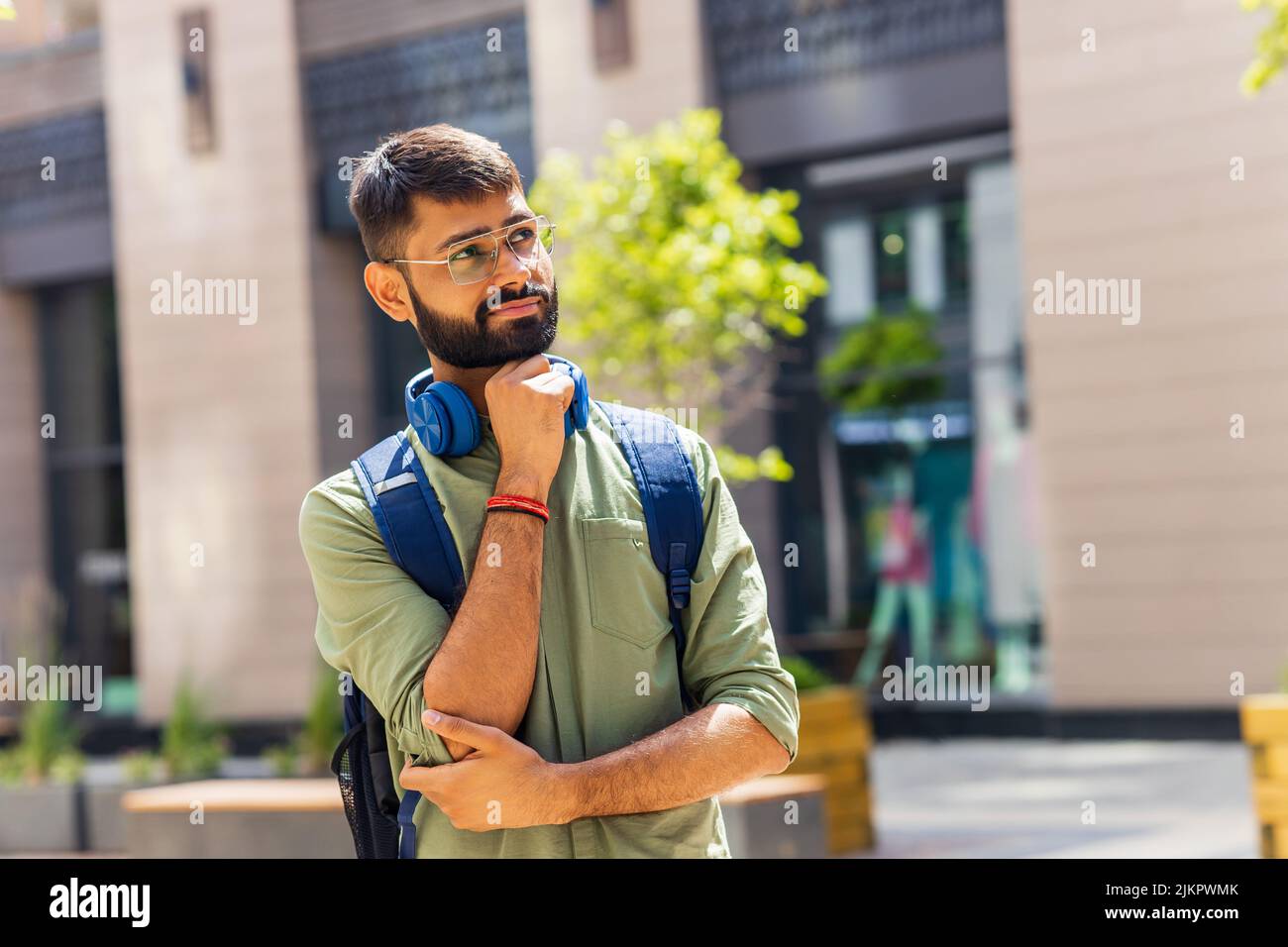 indischer Student mit blauem Headset und Rucksack, der an sonnigen Tagen nachdenklich aussieht Stockfoto