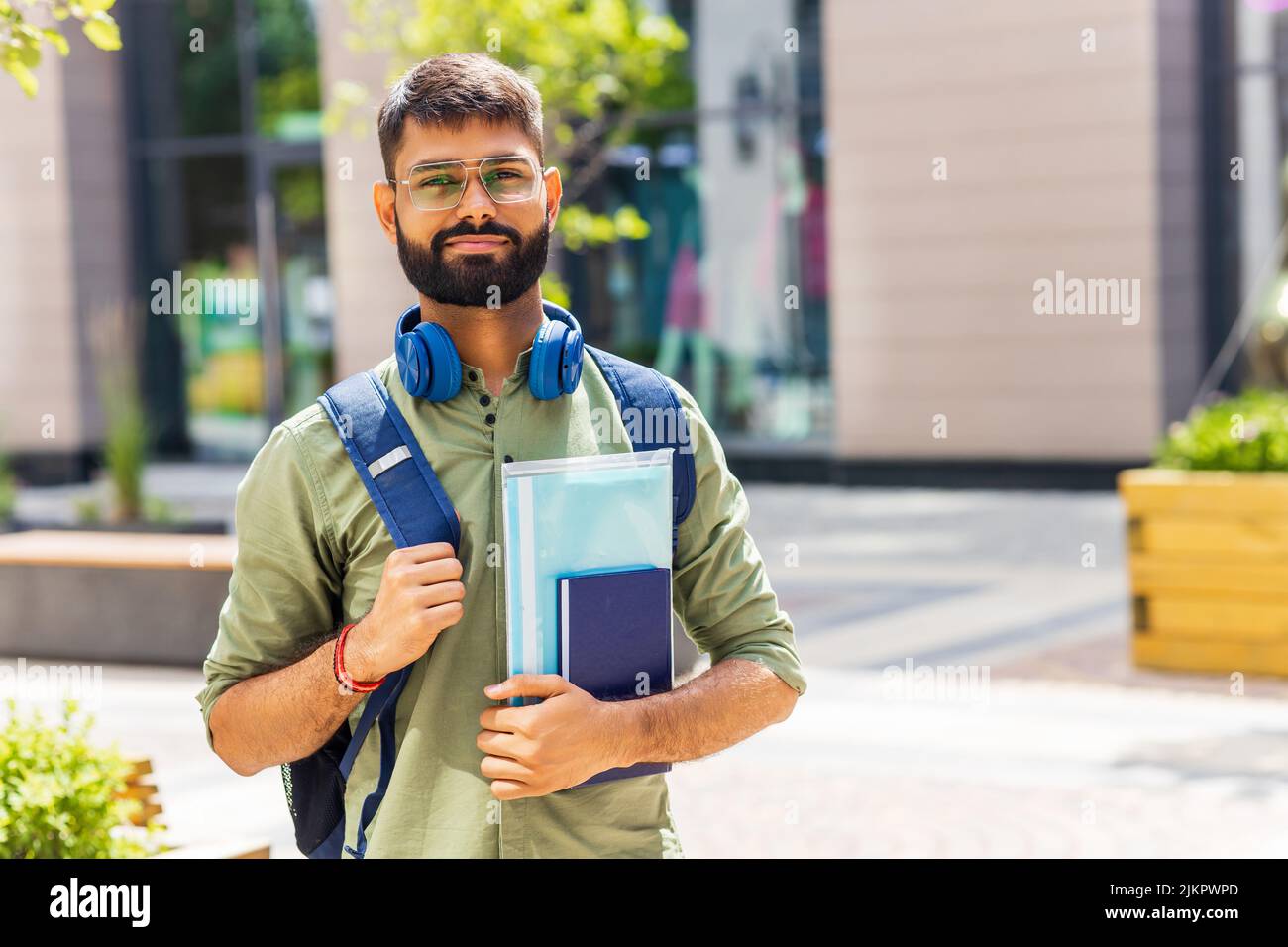 indischer Student mit blauem Headset und Rucksack, der an sonnigen Tagen Bücher hält Stockfoto