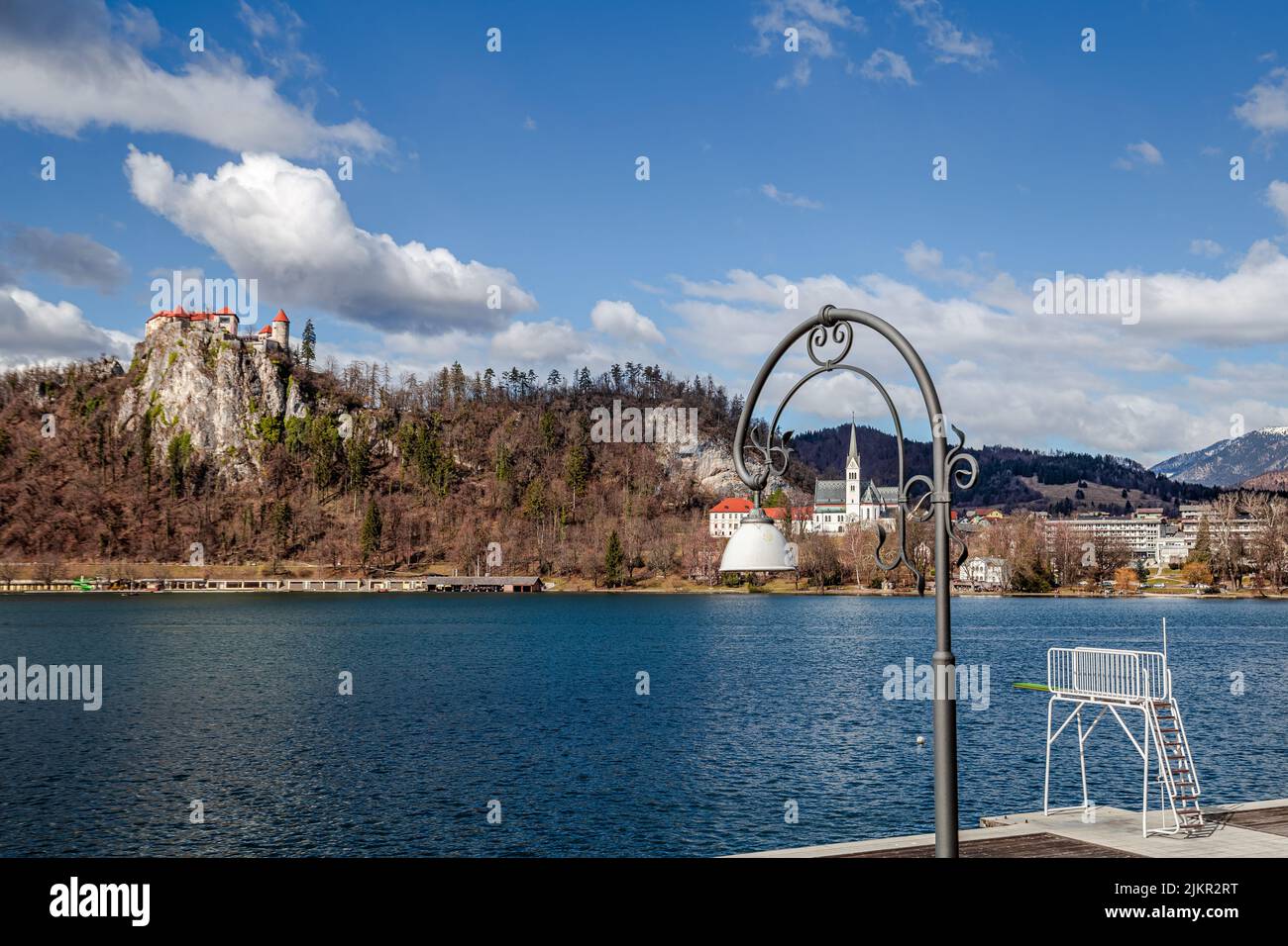 Bled, Slowenien - Lampenpfosten und Trampolin am Bleder See mit der Pfarrkirche St. Martin, der Burg von Bled und blauem Himmel und Wolken im Hintergrund bei einem sonnigen Wetter Stockfoto