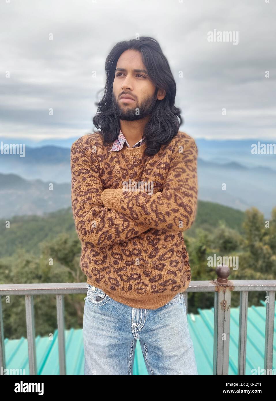 Vorderansicht eines südasiatischen jungen Mannes mit langen Haaren und Bart, der seitwärts schaut, während er mit gekreuzten Armen posiert, mit gelehnter Sicherheitsbarriere vor dem Hintergrund von Bergen steht Stockfoto